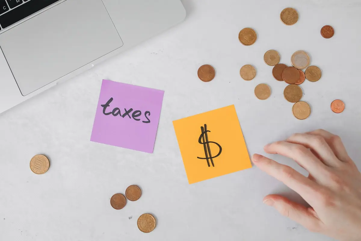Sticky notes reading 'taxes' and a dollar sign beside scattered coins and a laptop