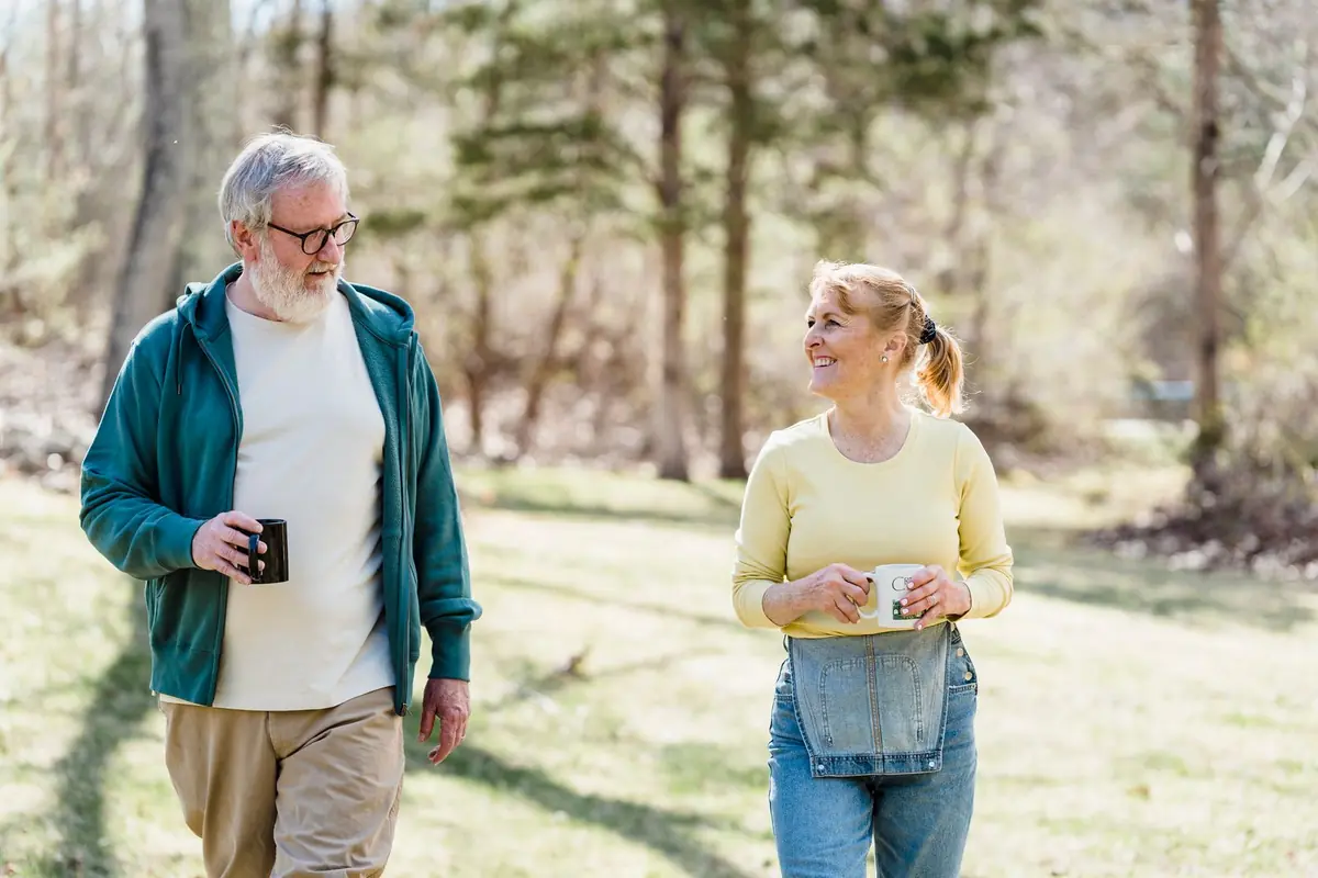 An older man and woman chatting as they walk outdoors among trees, each holding a hot drink